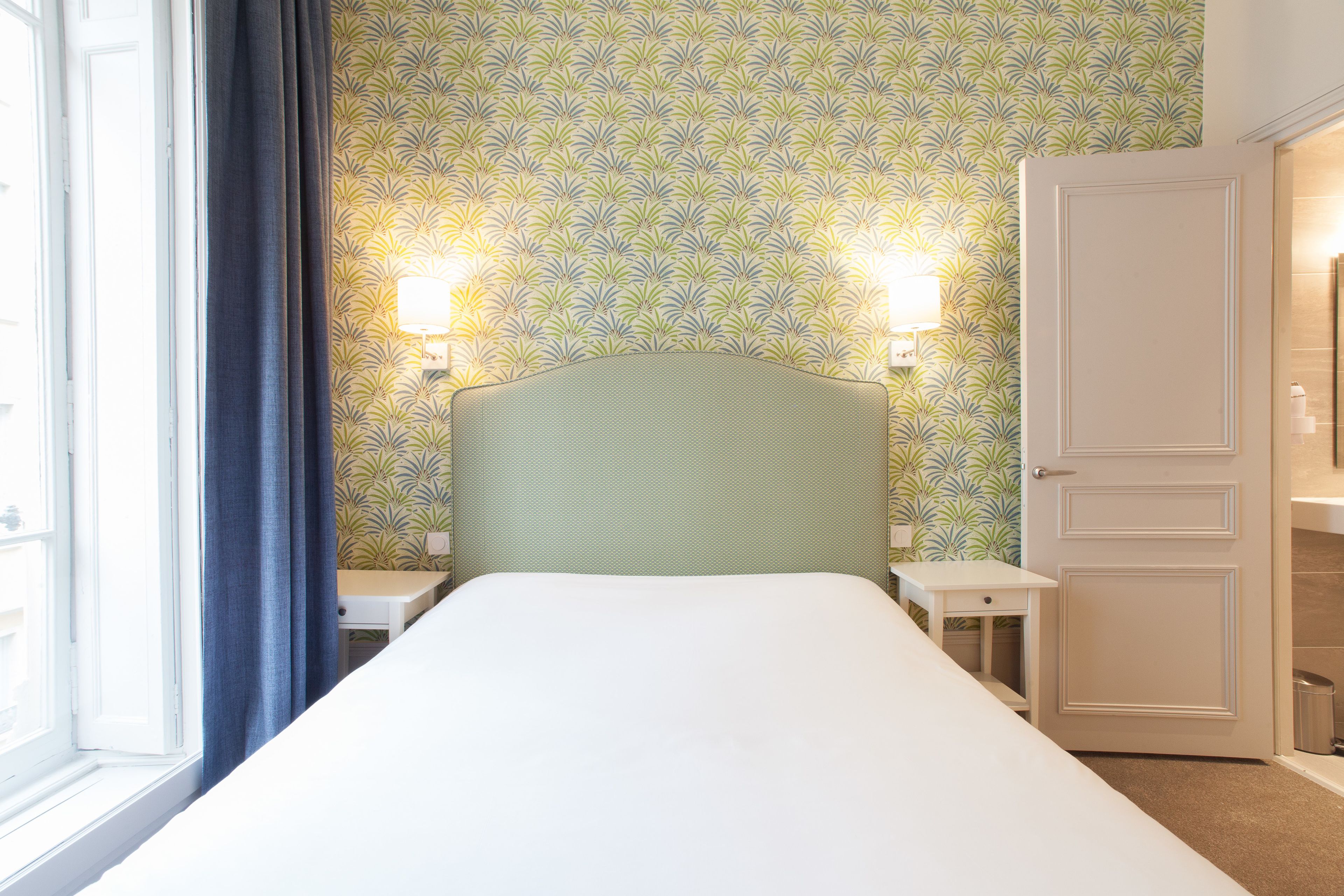 Une chambre d’hôtel comprenant un lit, deux tables de chevet, une tête de lit, une armoire et une fenêtre avec des rideaux.
