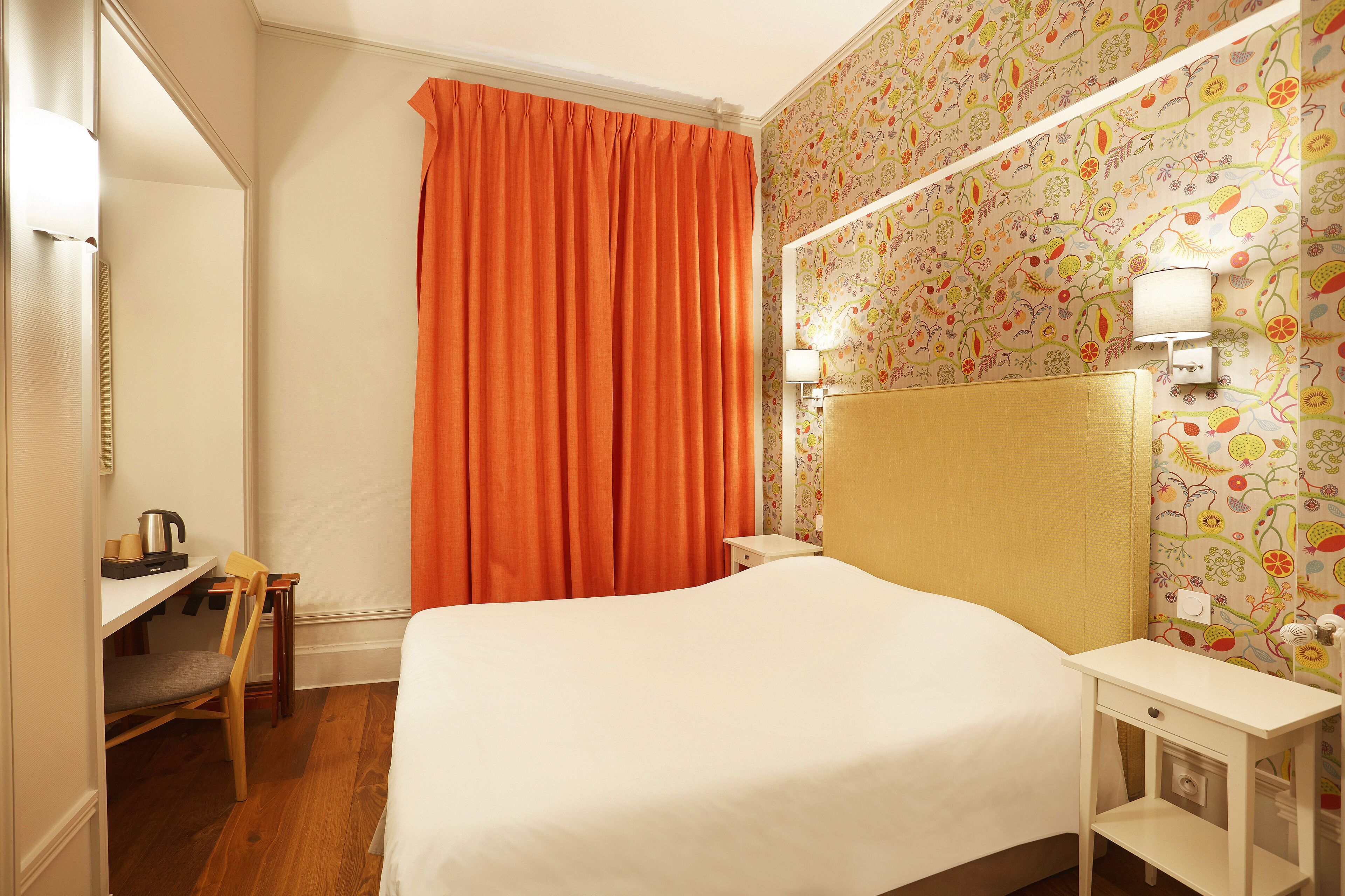 Une chambre d’hôtel comprenant un lit, un bureau avec une chaise, une table de chevet et une lampe fixée au mur.