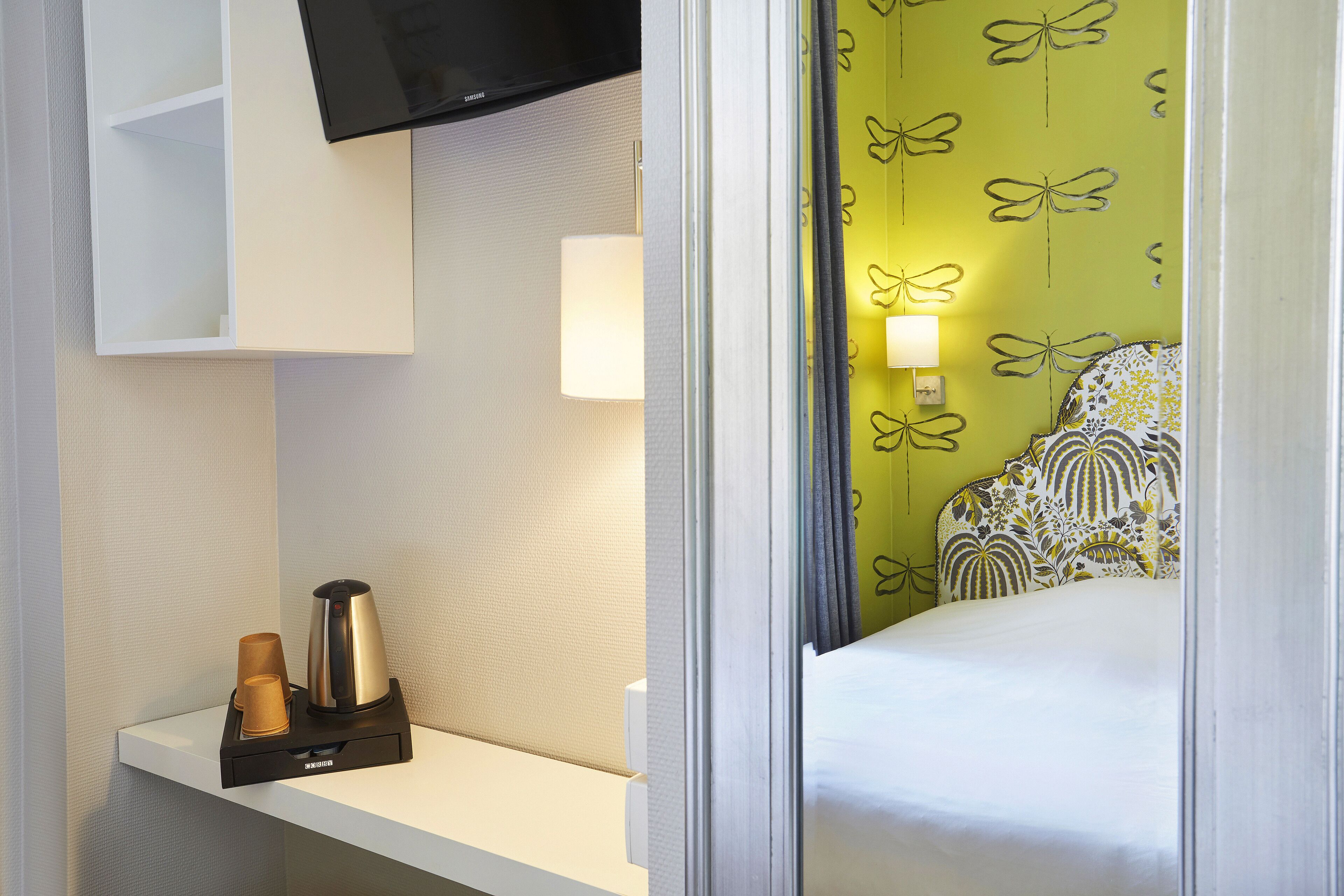 A hotel room with a bed, a TV mounted above a white shelf, a bedside lamp, and a wall with a butterfly pattern.