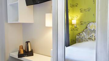 A hotel room with a bed, a TV mounted above a white shelf, a bedside lamp, and a wall with a butterfly pattern.