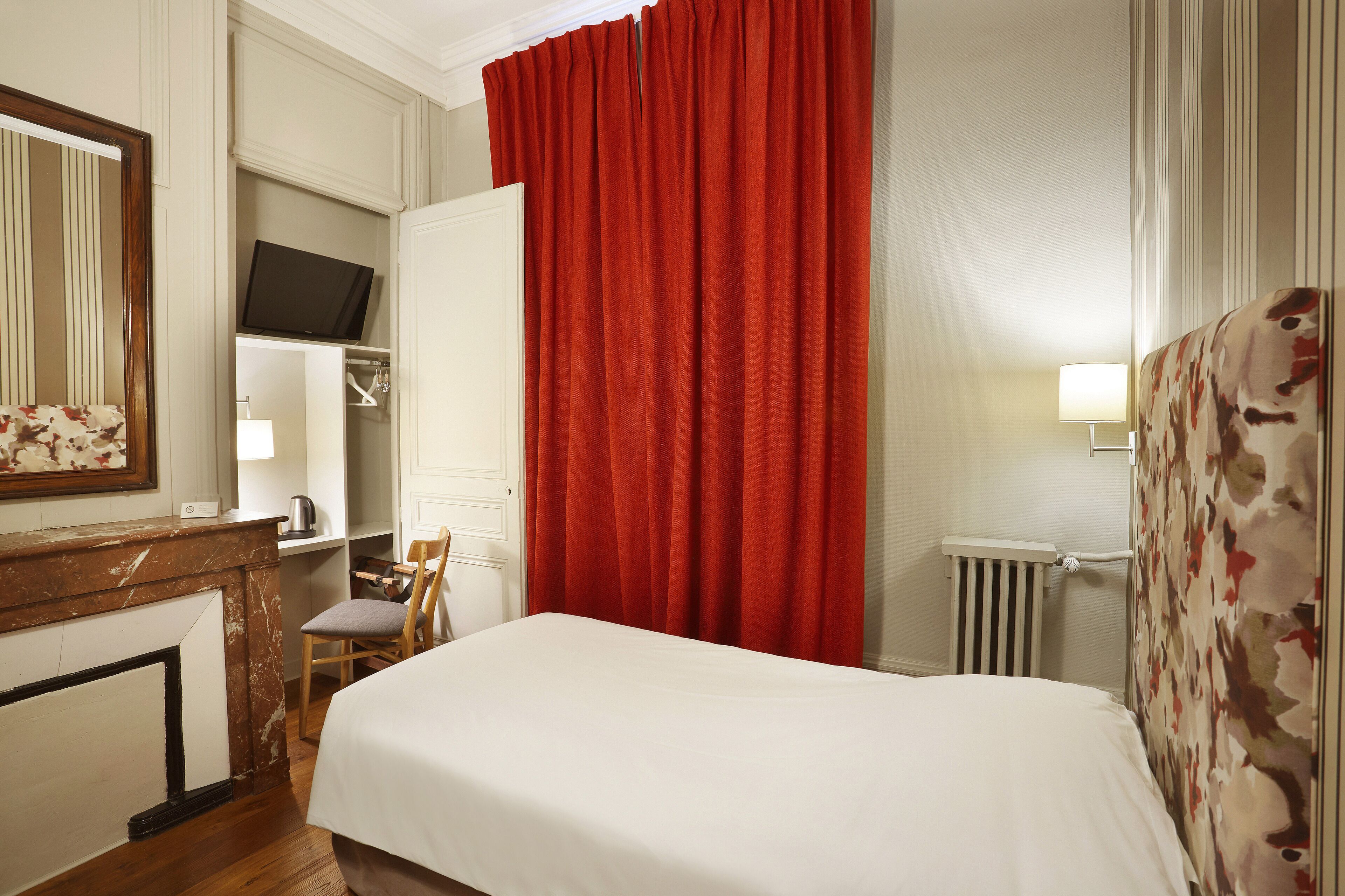 A hotel room with a bed, a chair, a desk with a mirror, a television, and a red curtain.