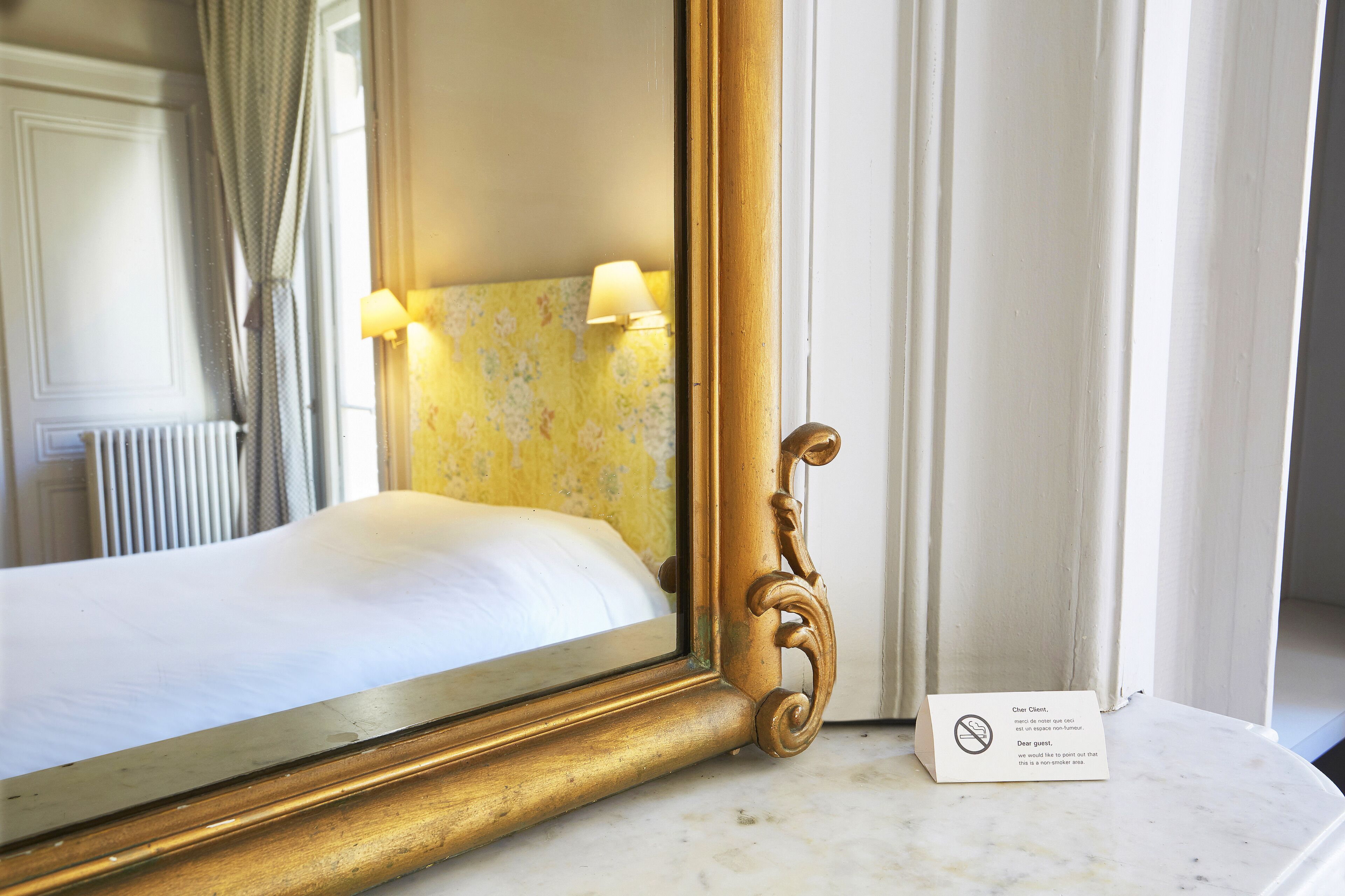Habitación de hotel con un espejo grande y ornamentado, una cama con ropa blanca, un cabecero estampado con motivos florales y un letrero que indica que no está permitido pintar.