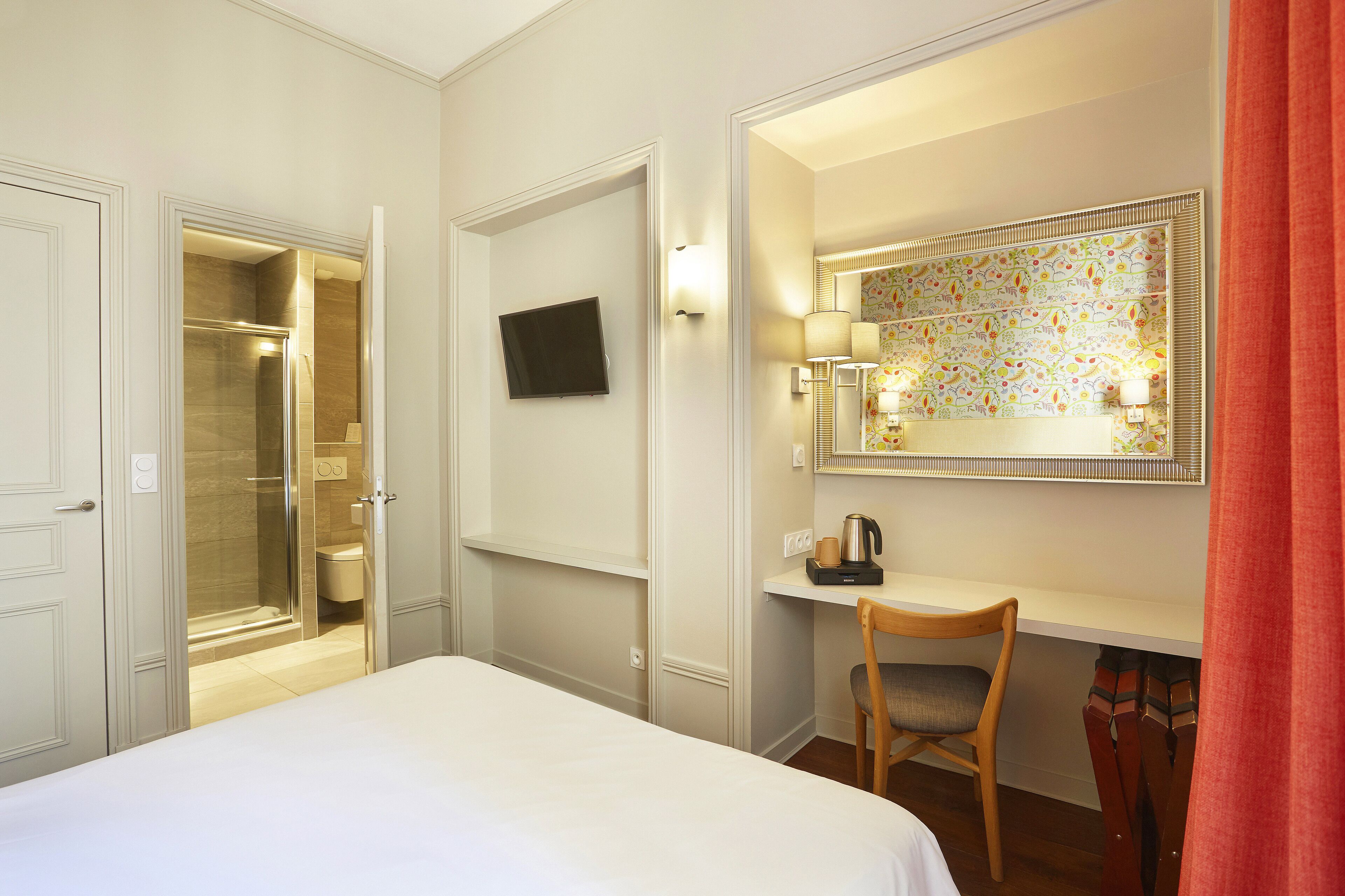 A hotel room with a bed, a desk with a chair, a shower, and a television on the wall.