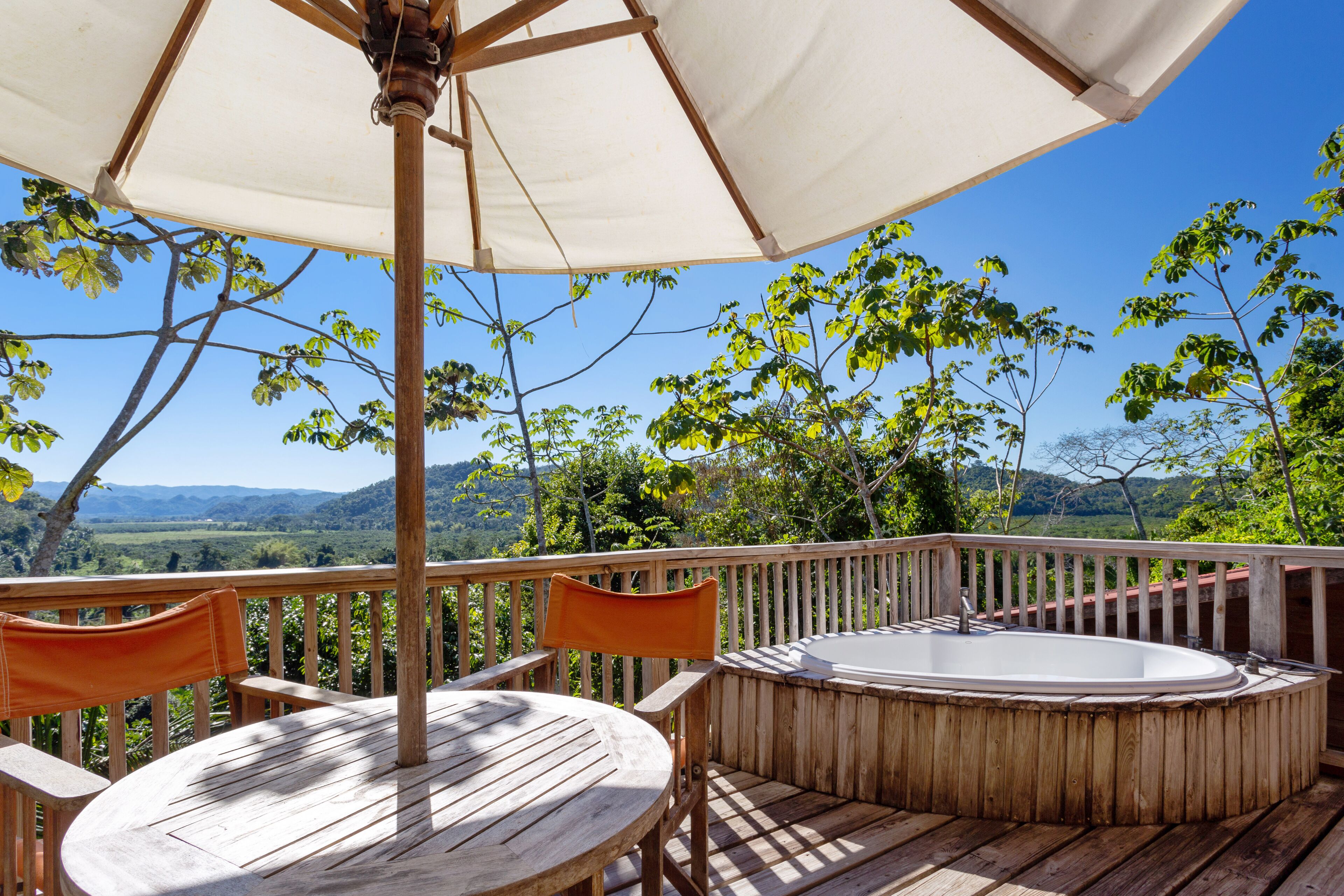Una terrazza in legno con vasca idromassaggio, tavolo e sedie sotto un grande ombrellone.