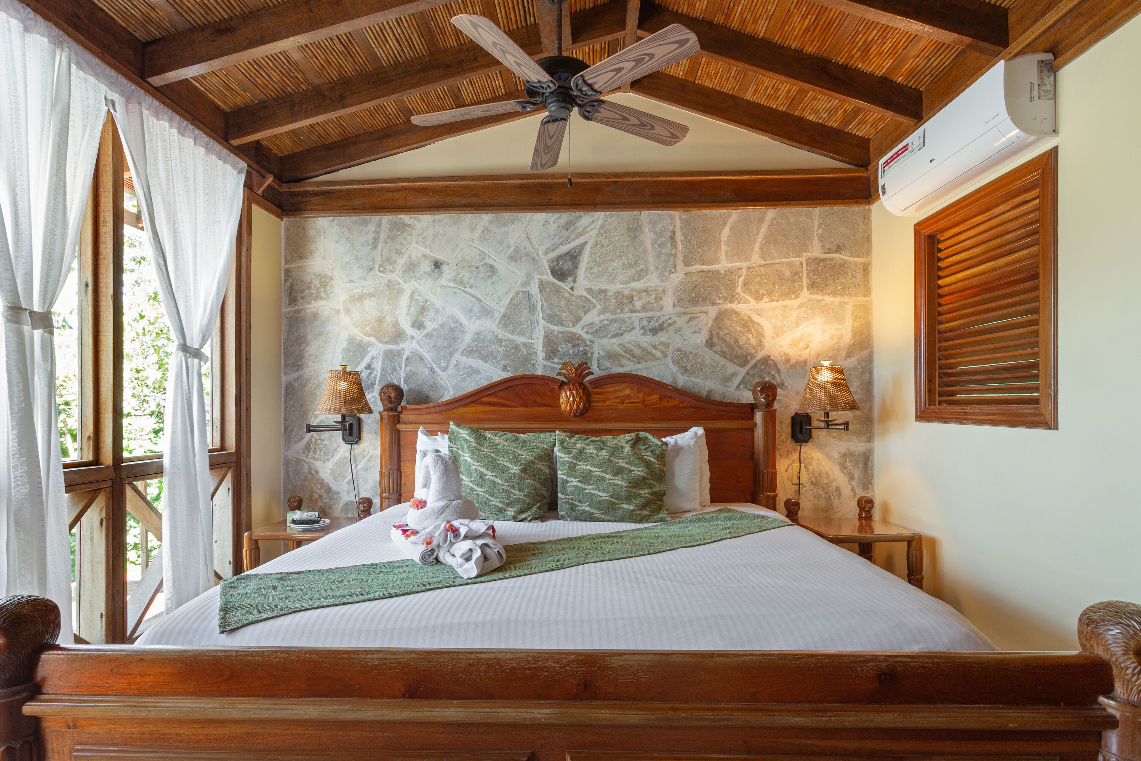 A bedroom with a stone wall, wooden ceiling beams, a bed with green and white bedding, two wall-mounted lamps, and a ceiling fan.