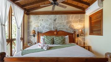 Un dormitorio con pared de piedra, vigas de madera en el techo, cama con ropa de cama verde y blanca, dos lámparas de pared y un ventilador de techo.