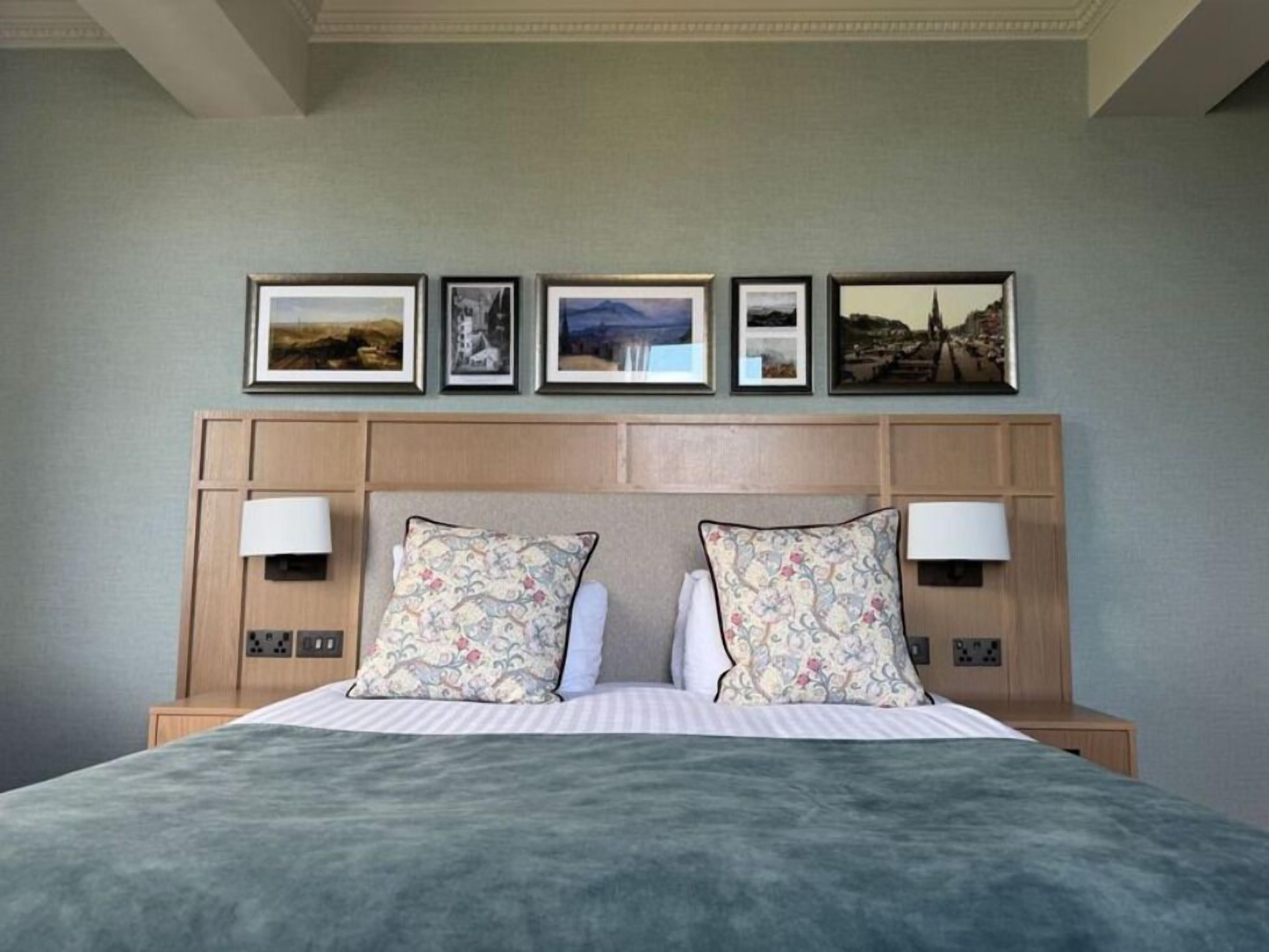 Une chambre d’hôtel avec un lit, deux lampes de chevet, une tête de lit ornée de tableaux encadrés et un mur aux couleurs neutres.