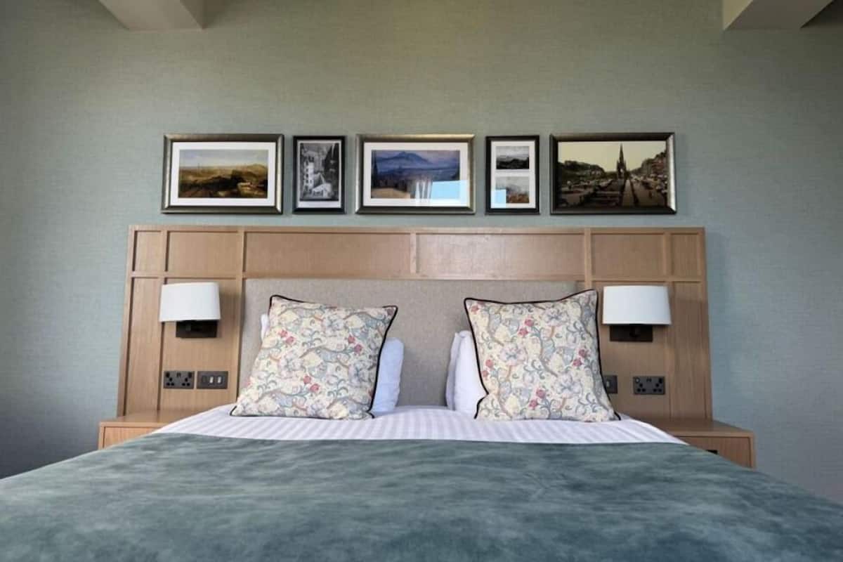 A hotel room with a bed, two bedside lamps, a headboard with framed pictures, and a neutral-colored wall.