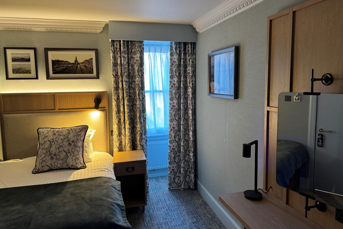 Une chambre d’hôtel comprenant un lit, une table de chevet, une lampe, des rideaux, des tableaux encadrés et un téléviseur.