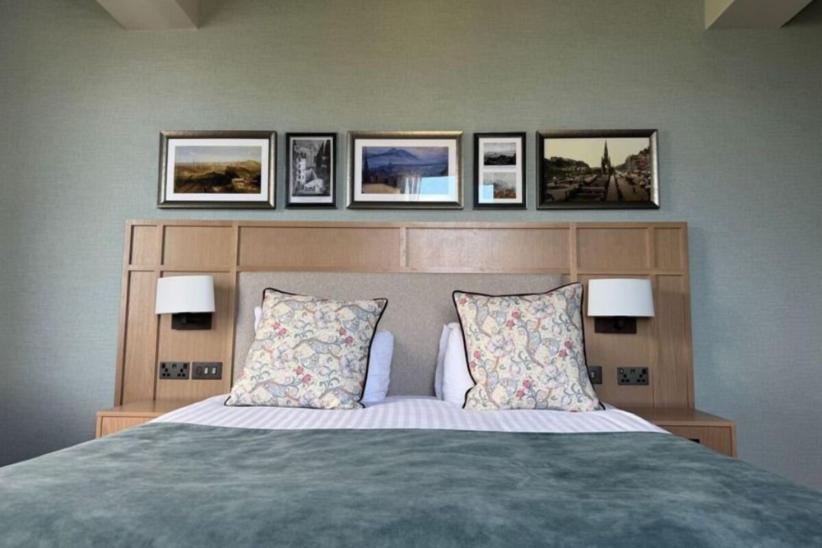 A hotel room with a bed, two bedside lamps, a headboard with framed pictures, and a neutral-colored wall.