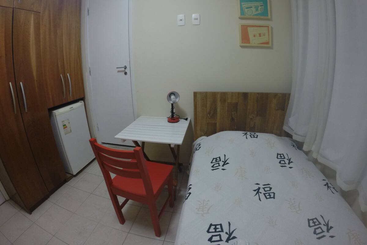 Ein kleines Zimmer mit einem Bett, einem Schreibtisch, einem Stuhl, einem Kleiderschrank und einem kleinen Kühlschrank.