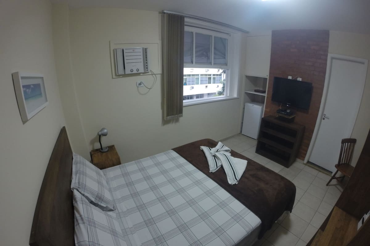 Ein Schlafzimmer mit Bett, Fenster, Fernseher und einer kleinen Küchenzeile.
