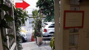 Ein schmaler Eingang mit einem roten Pfeil, der nach rechts zeigt, führt zu einer Straße mit parkenden Autos und einem weißen Auto im Hintergrund.