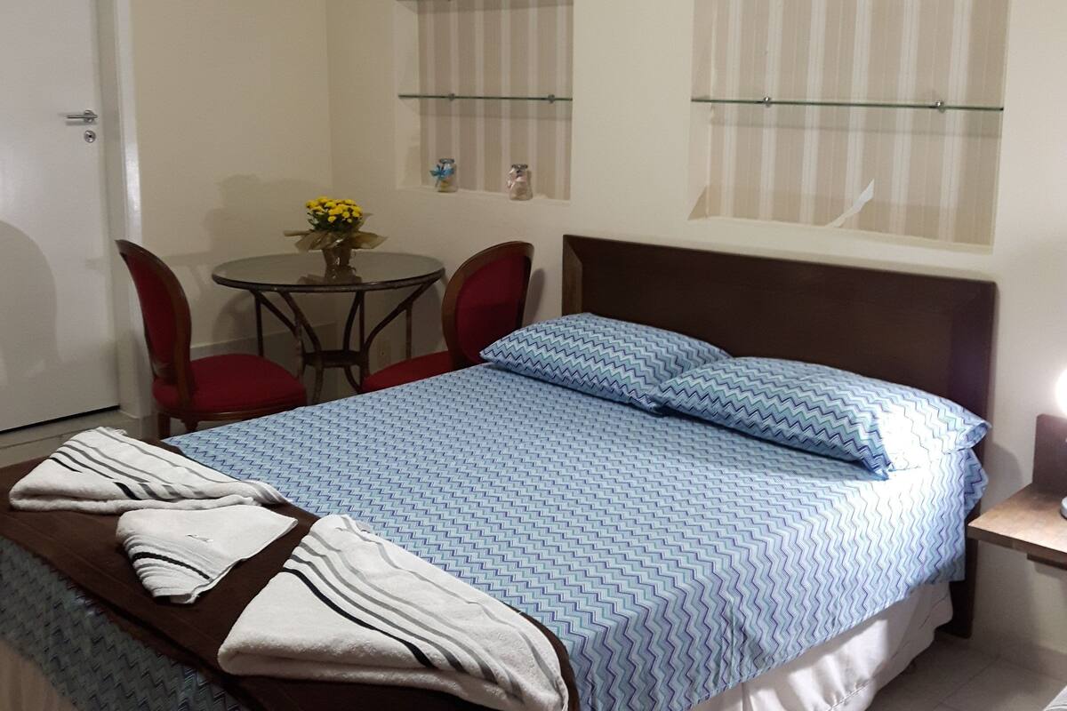Ein Schlafzimmer mit einem Bett, einem kleinen Tisch mit Stühlen und zwei Fenstern mit gestreiftem Vorhang.