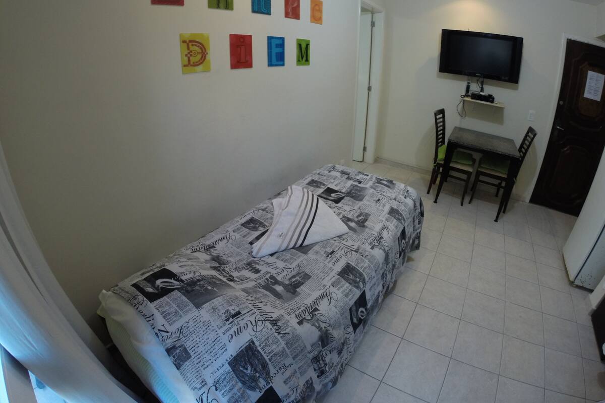 Ein kleiner, ordentlicher Raum mit einem Einzelbett, einem Schreibtisch mit Stuhl, einem an der Wand befestigten Fernseher und einer Tür zu einem weiteren Raum.