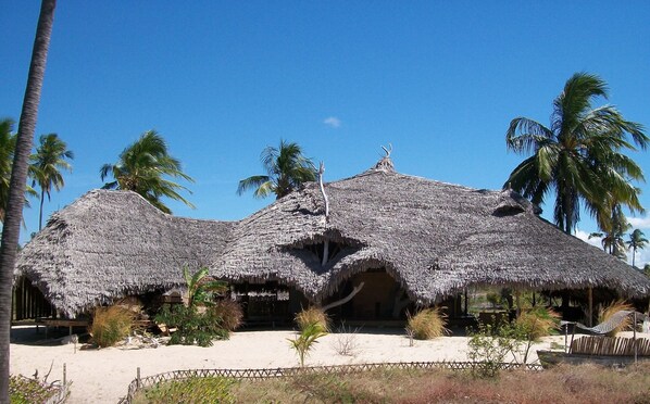 Estructura de techo de paja con varias habitaciones, rodeada de palmeras y una zona arenosa.