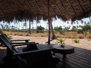 Una terraza de madera con sillones de descanso y una pequeña mesa, bajo techo de paja, con vista a una playa de palmeras y pasto seco.
