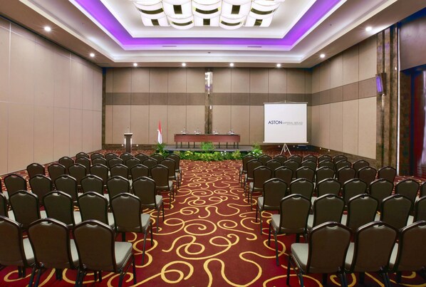 A conference room with rows of chairs facing a podium and a screen displaying "ASTON IMPERIAL EXCHANGE".