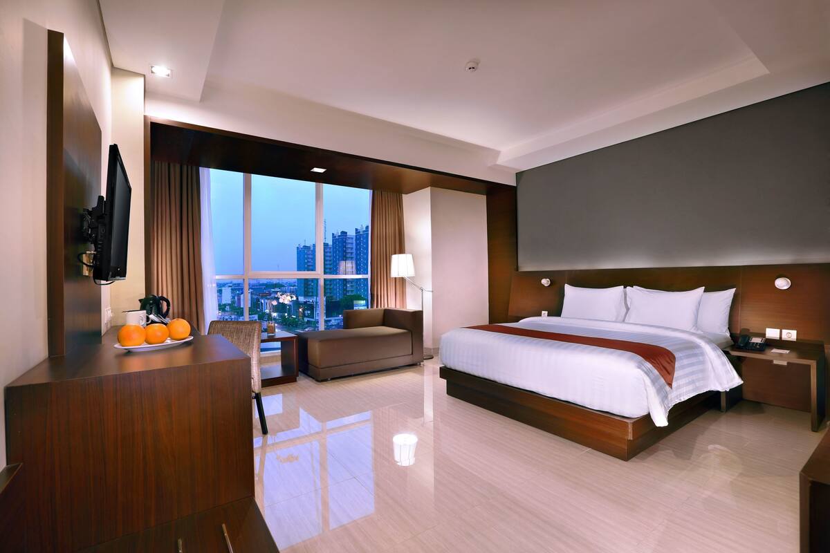 Une chambre d’hôtel moderne avec un grand lit, un canapé, un bureau orné d’oranges, une télévision et une vue sur la ville.