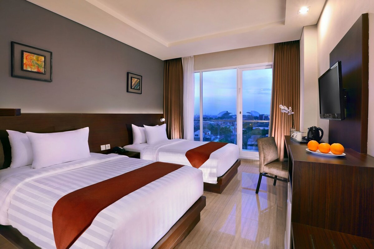 Habitación de hotel con dos camas, un escritorio con silla, televisión y vista panorámica de la ciudad.