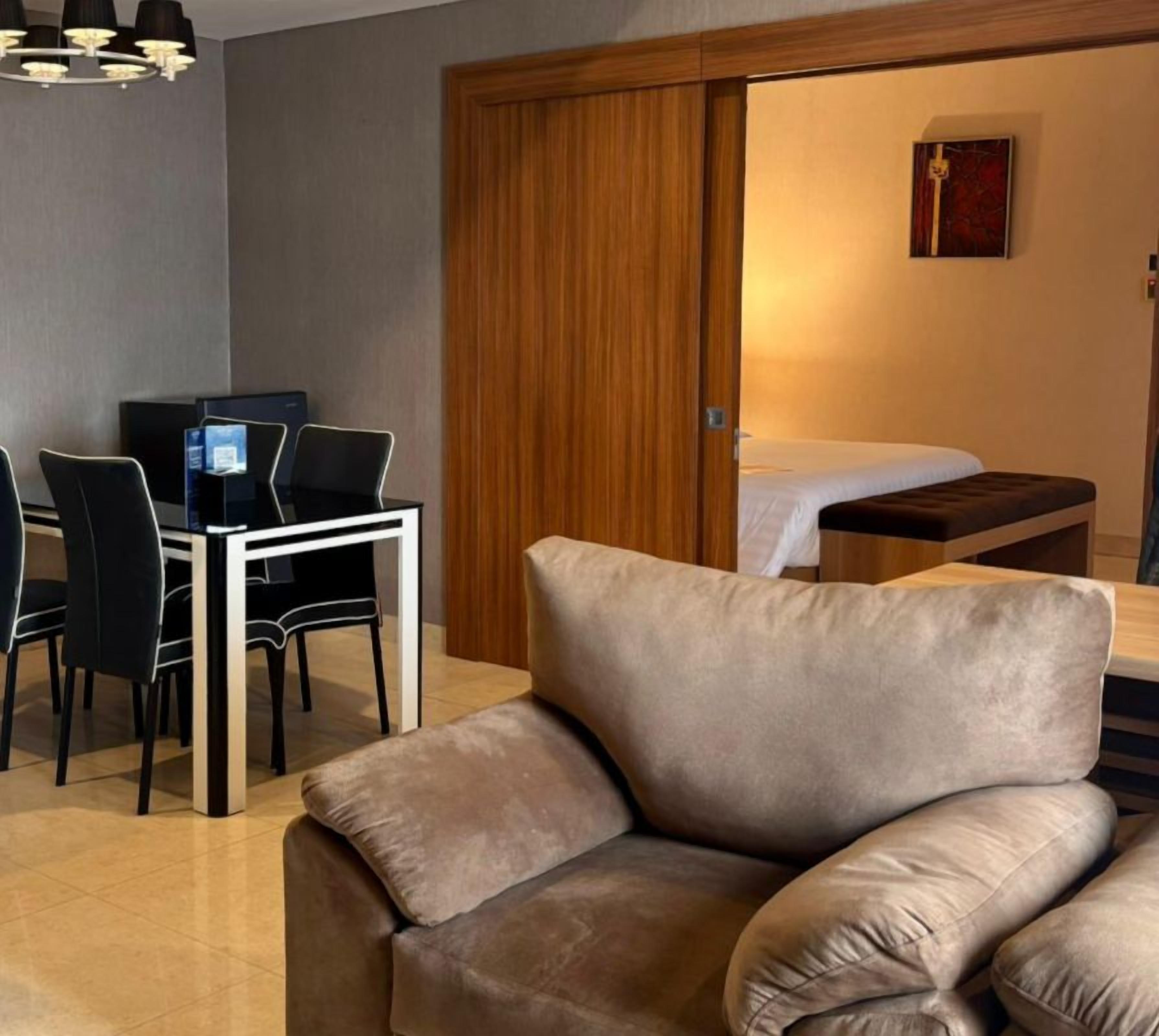 A modern hotel room with a dining area, a sofa, a bed, and a wooden door.