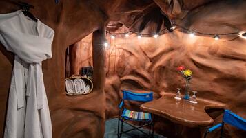 A room with a rustic, cave-like interior, featuring a wooden table with a vase of flowers, a blue chair, and a rack of white robes.
