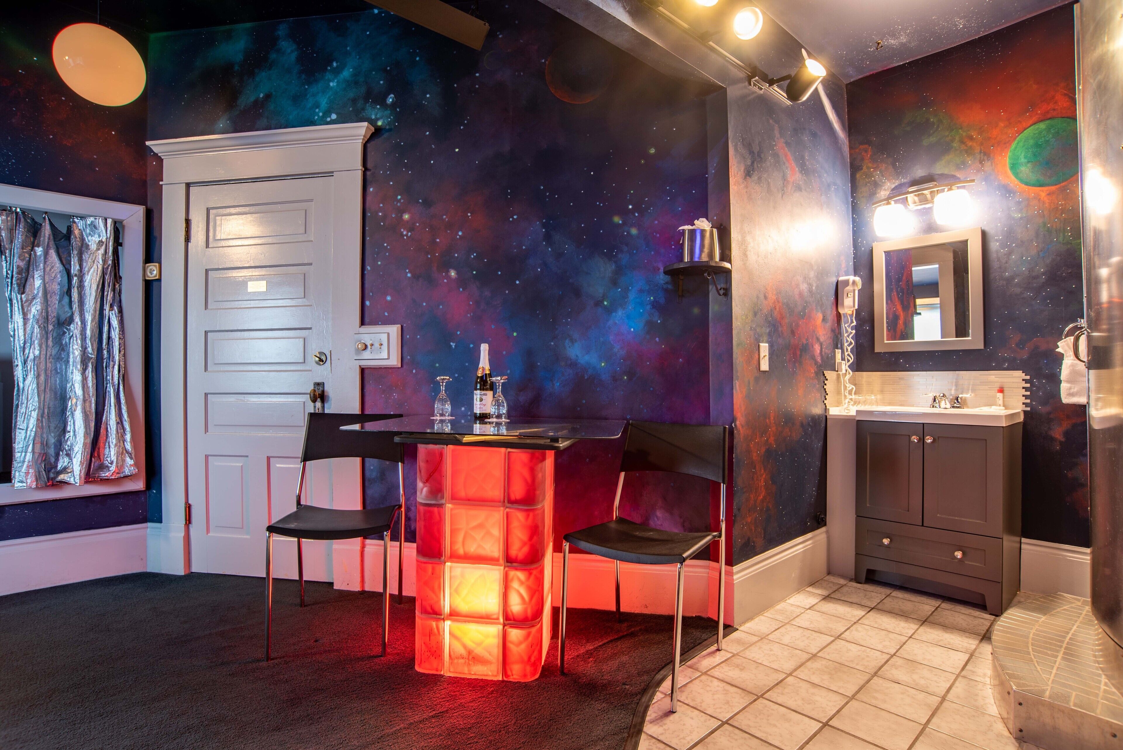 Een badkamer met een wanddecoratie in een galaxiethema, een wastafel met spiegel, en een klein tafeltje met stoel.