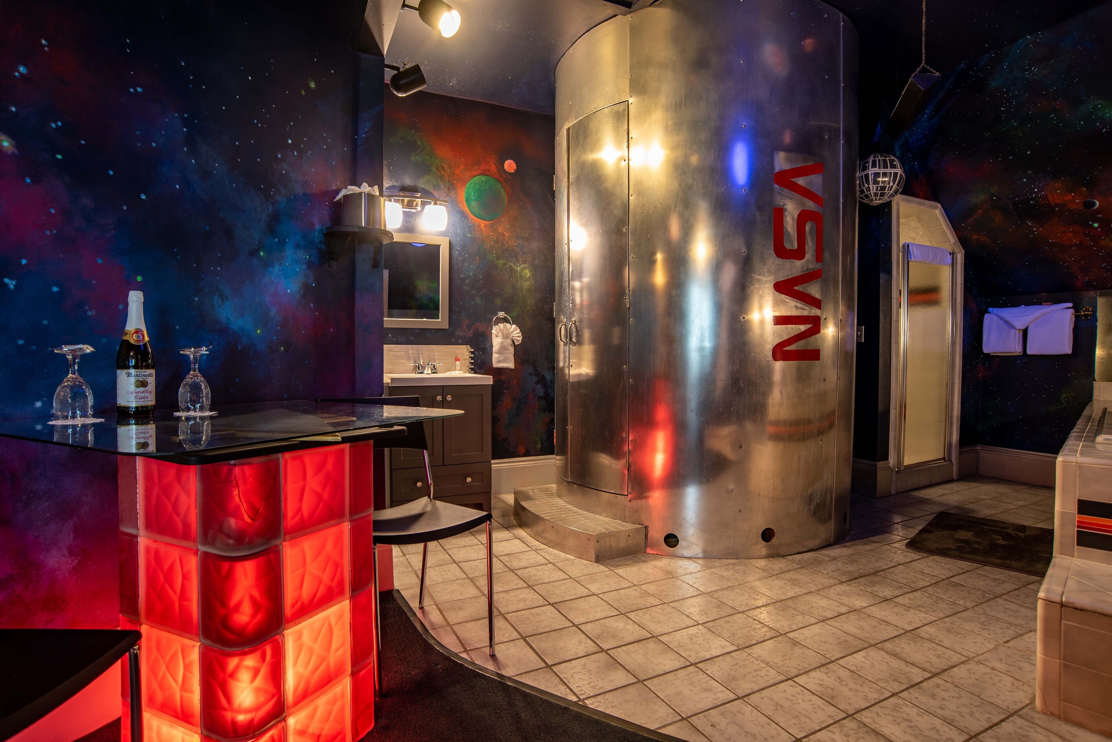 A space-themed room with a metallic cylindrical structure labeled 'NASA', a bar area with glasses, and a bathroom with a space mural.