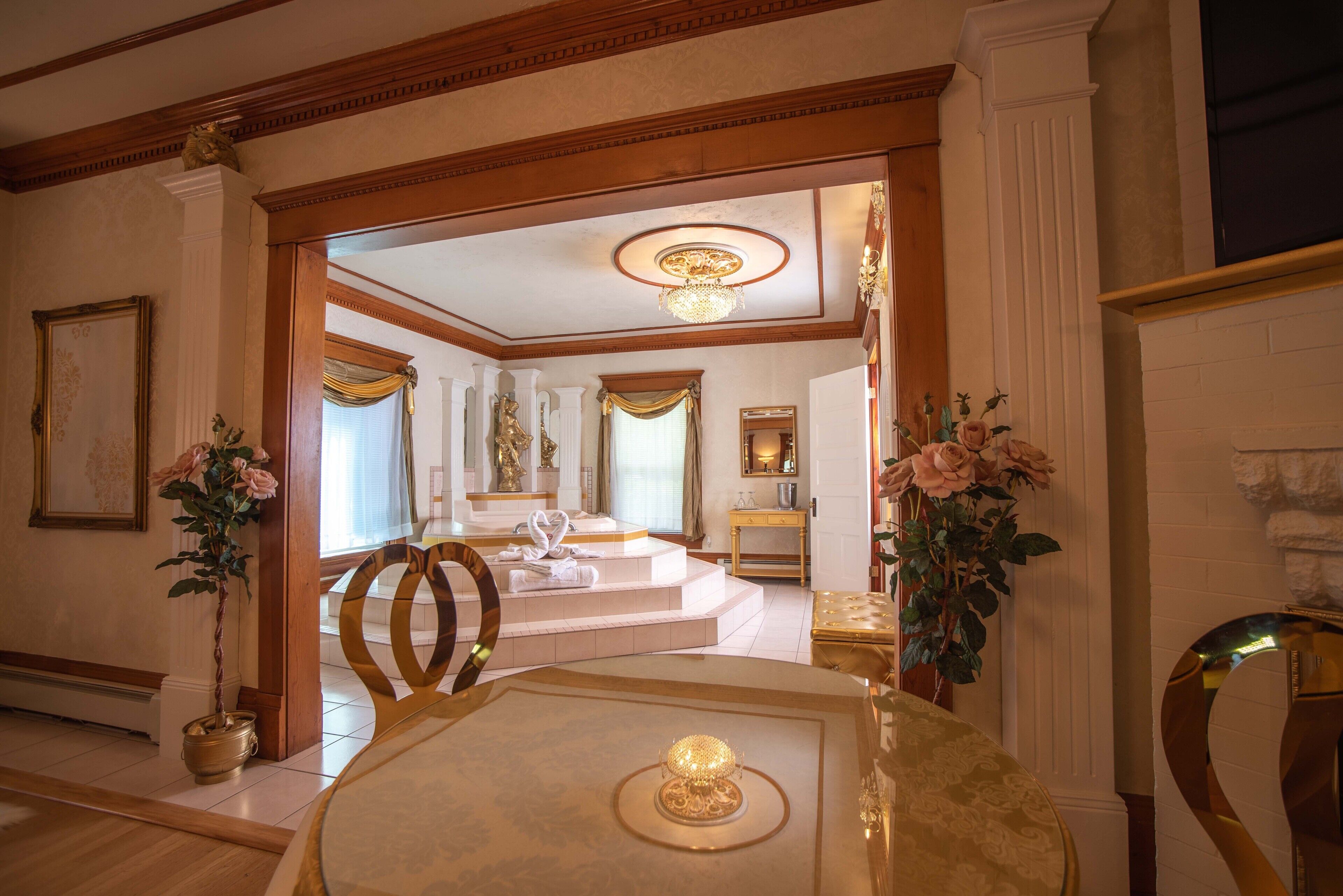 A luxurious hotel room with a large bed, a sitting area with a table and chairs, and a view of the adjacent room.