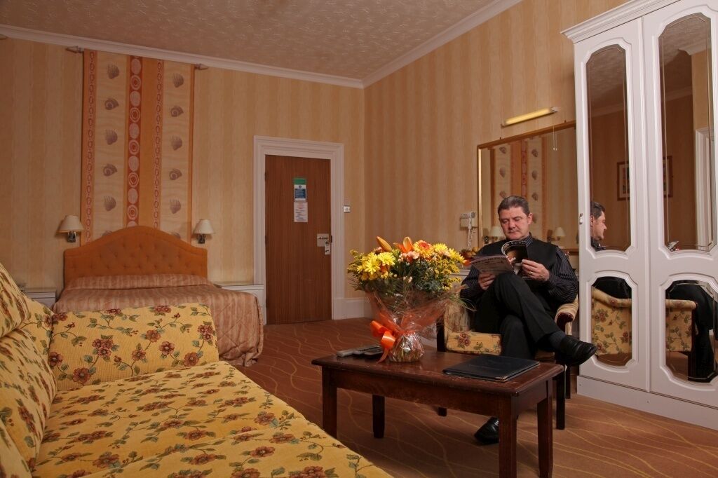 A hotel room with a bed, a sofa, a table with a vase of flowers, and a mirror cabinet.