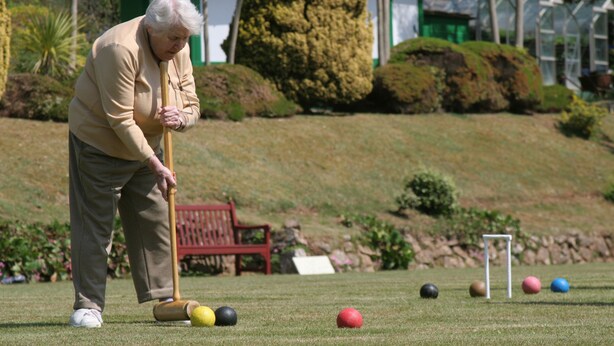 An elderly person playing croquet on a well-maintained lawn with a red bench and a house in the background.