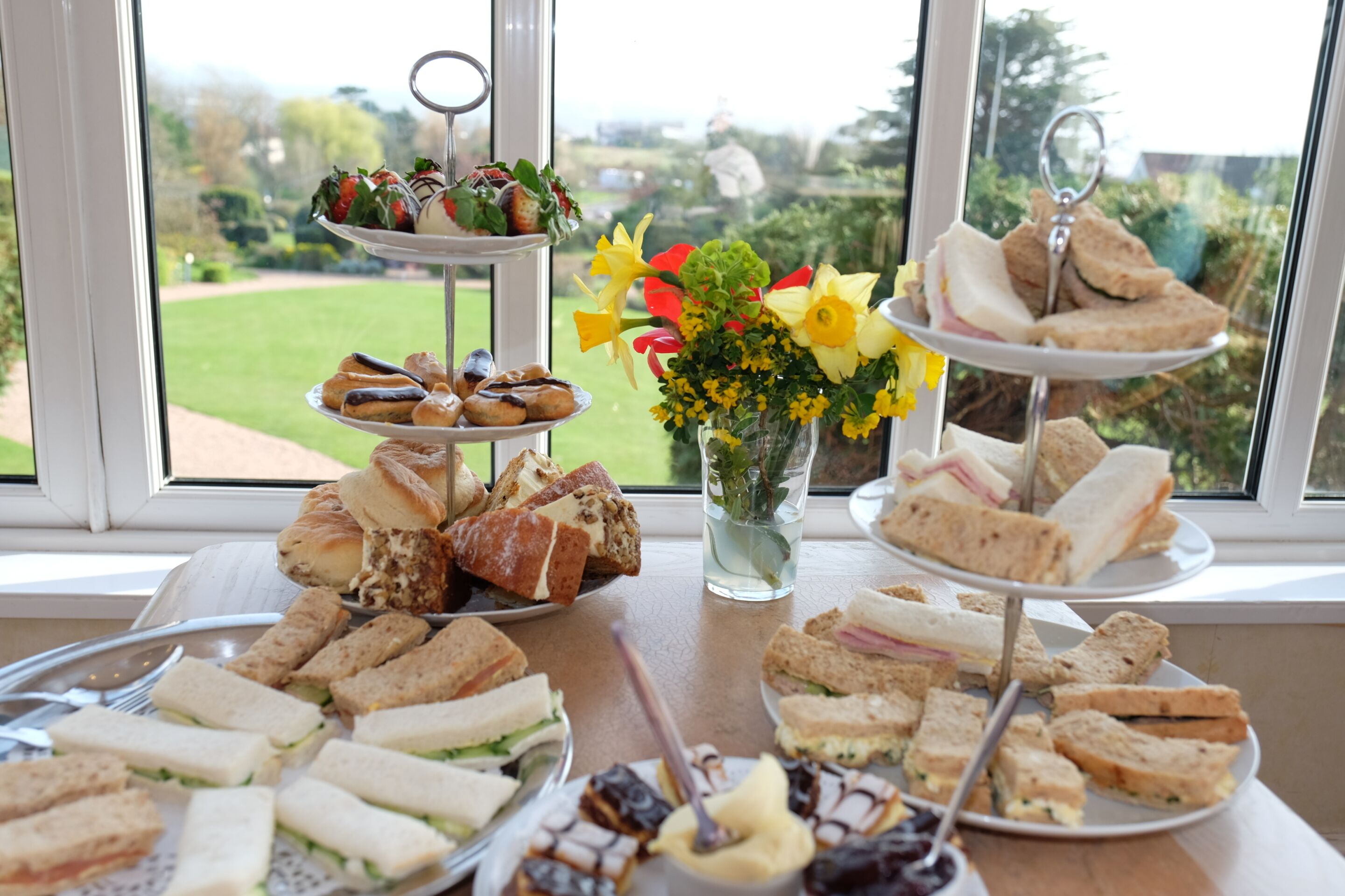 A table with various sandwiches and pastries, a vase with flowers, and a window view.