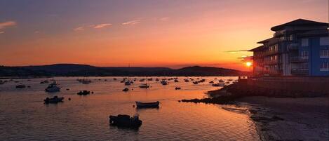 Escena costera con barcos en aguas tranquilas, un edificio con balcones y un atardecer al fondo.