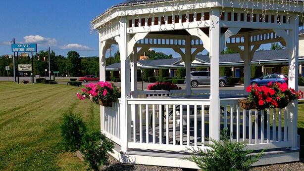 A gazebo with a white structure and a roof, surrounded by a white picket fence, with pink flowers in planters. - WYE Motor Lodge Duncansville - Altoona (Hollidaysburg)