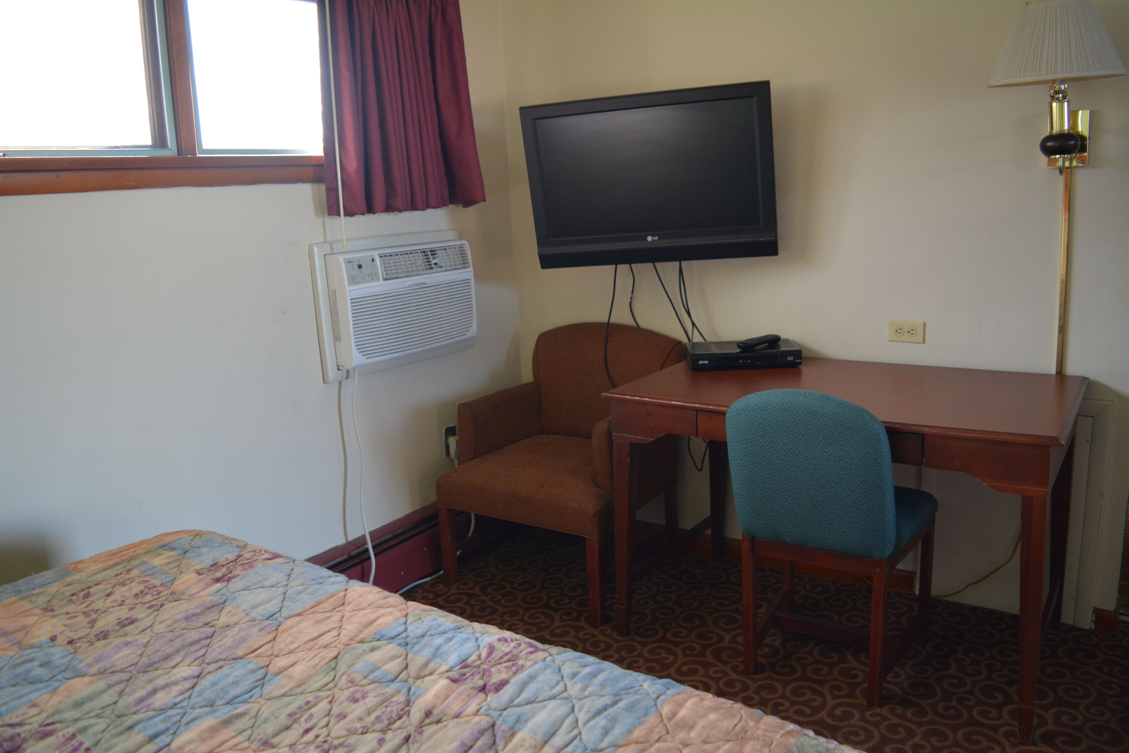A hotel room with a bed, a desk with a chair, a television, an air conditioner, and a lamp.