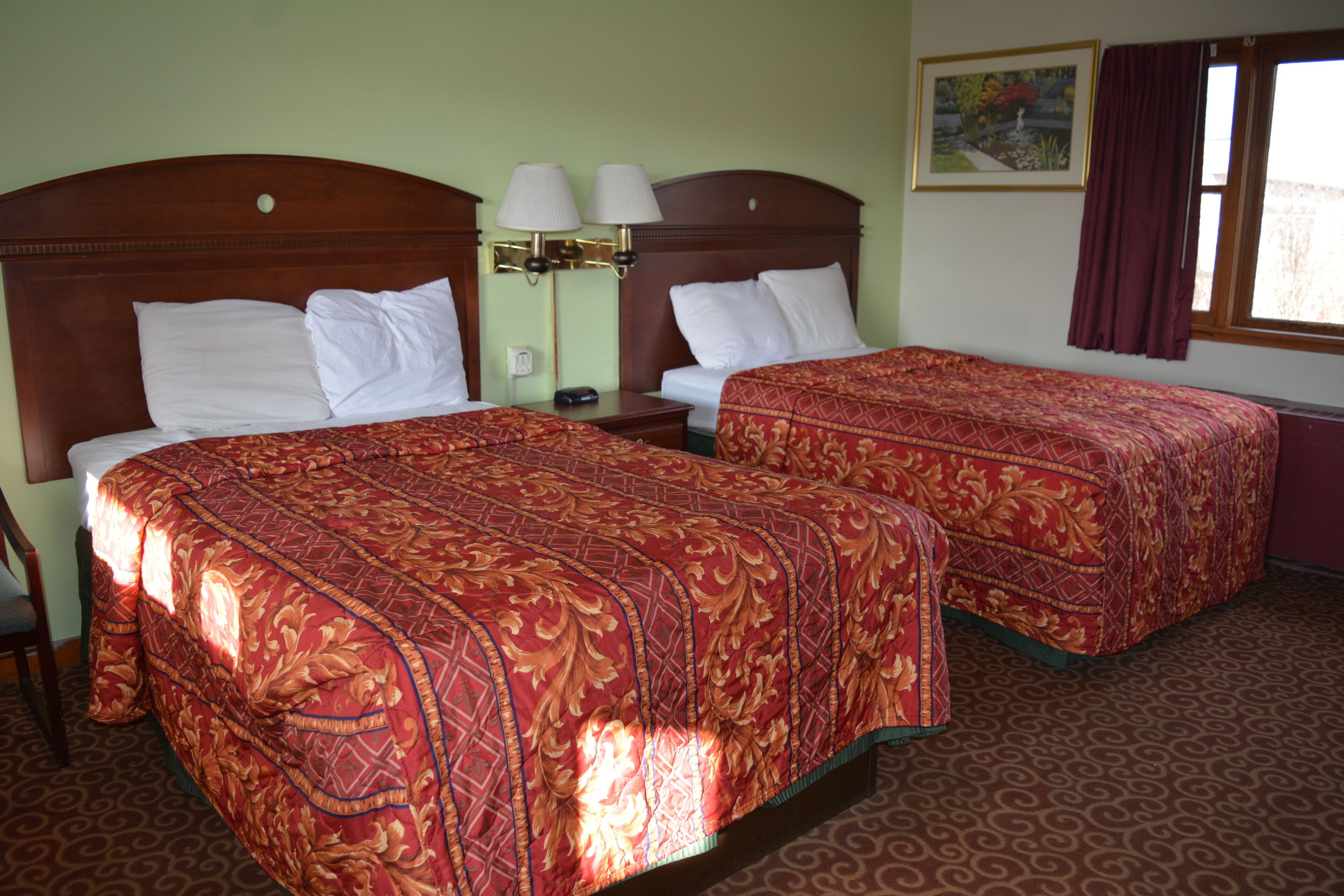 Habitación de hotel con dos camas, cada una con cabecero de madera, una mesita de noche con lámparas y un ventanal con cortinas.