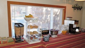 A breakfast buffet setup with a variety of food items and beverages, a coffee machine, and a window with blinds. - Kokopelli Inn (Bluff)
