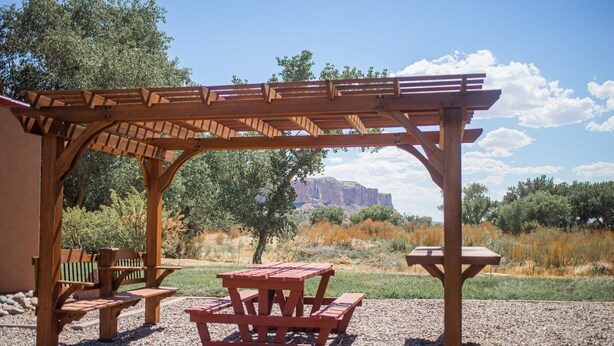 A wooden pergola with picnic tables and benches, set against a backdrop of trees and a mountain. - Kokopelli Inn (Bluff)