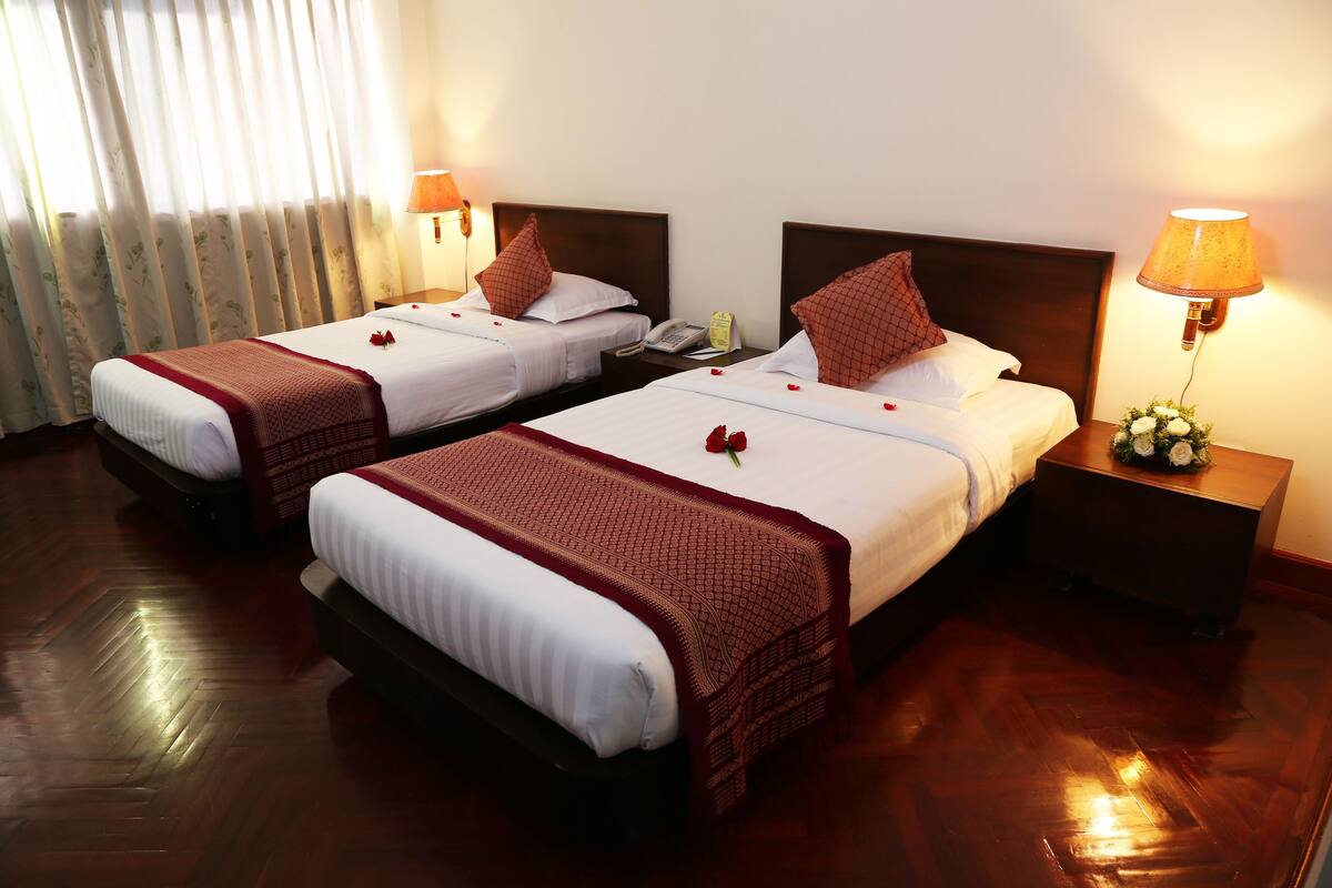 A hotel room with two beds, wooden flooring, and a nightstand with a lamp.