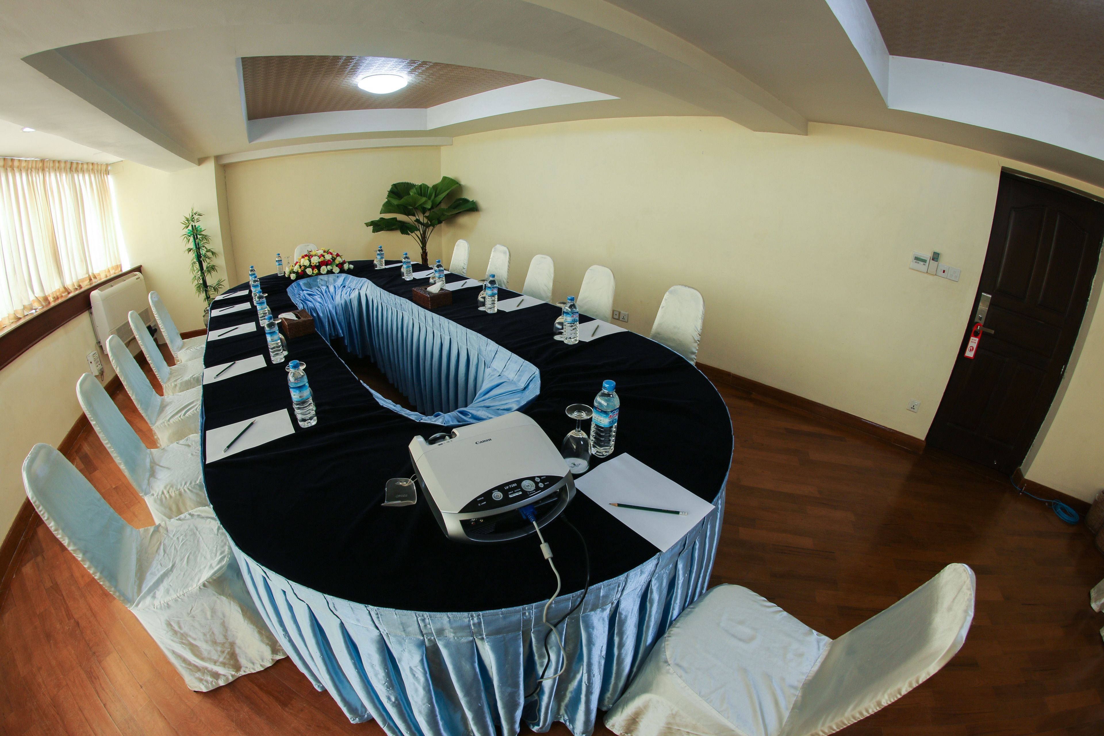 Un salón de conferencias con una larga mesa, sillas blancas, un proyector, botellas de agua y una planta en maceta.