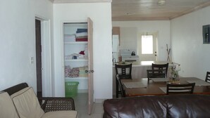 Una sala de estar con sofá, una mesa de comedor con sillas, una zona de cocina con estufa y refrigerador, y un cuarto de lavado con estantes y una cesta.