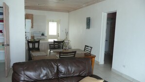 Una sala de estar con un sofá de cuero marrón, una mesa de comedor con sillas, y una cocina al fondo.