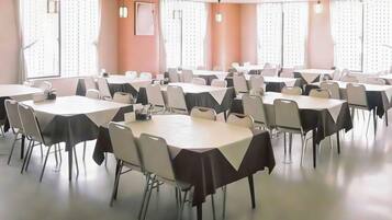 A conference room with multiple tables and chairs, large windows with blinds, and ceiling lights.