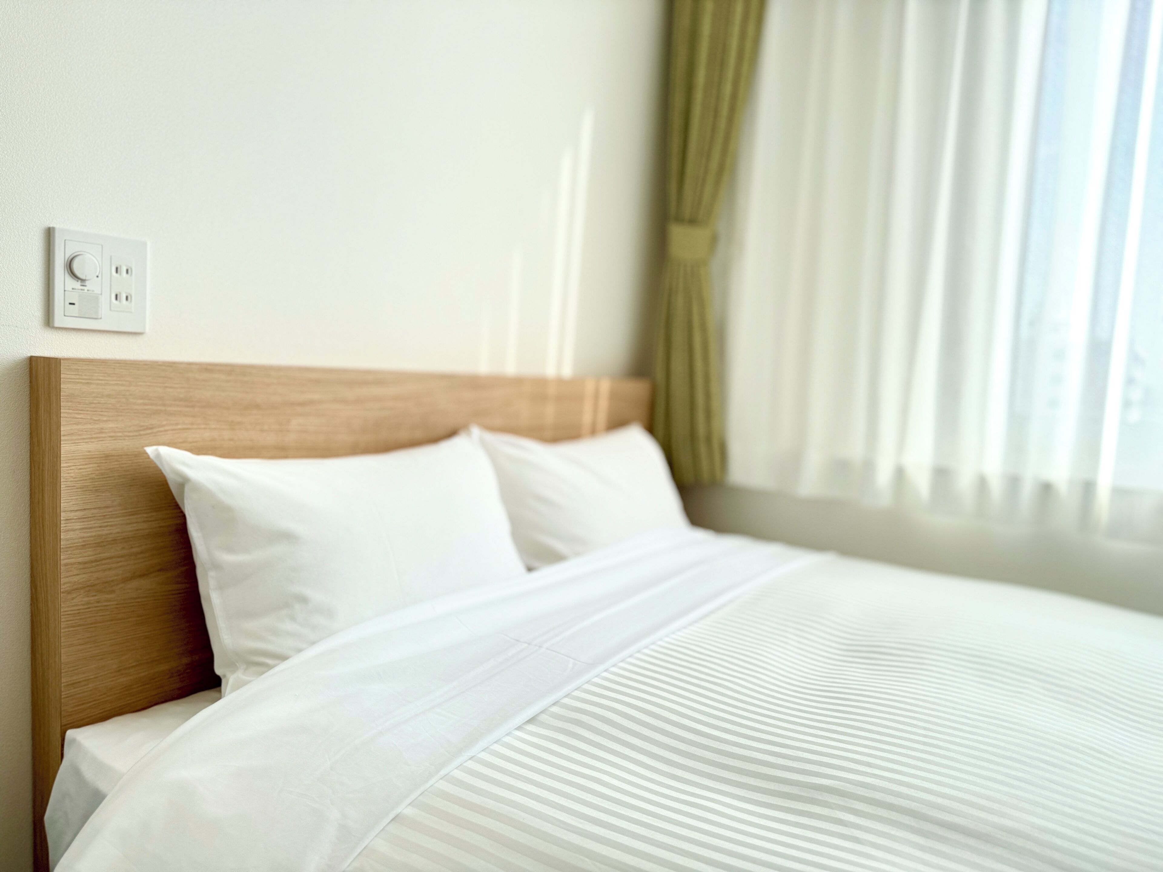 A neatly made bed with white linens, a wooden headboard, and a wall-mounted light switch.