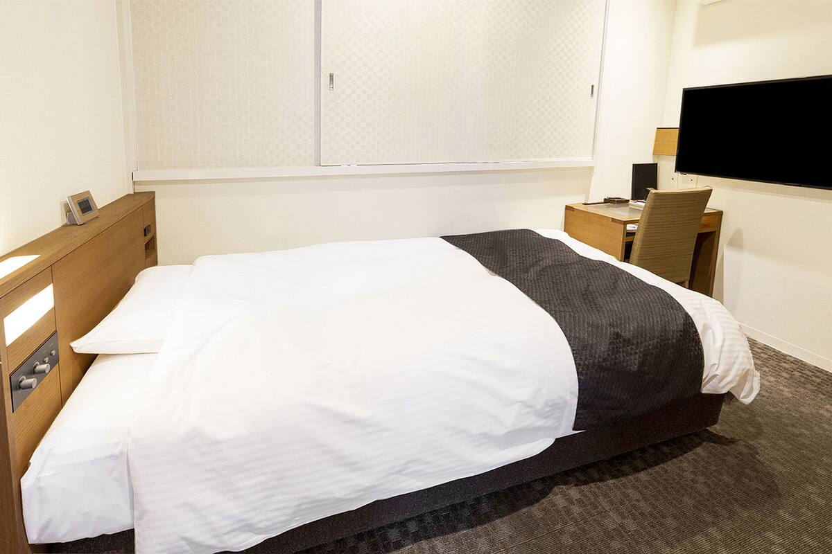 Quarto de hotel com cama, escrivaninha, cadeira, televisão e guarda-roupa.