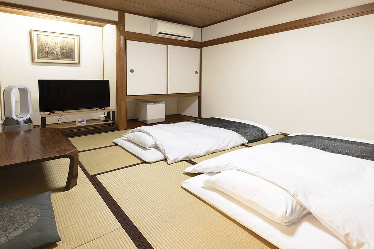 Quarto de hotel com piso de tatame, uma cama, uma escrivaninha, uma televisão e um ar condicionado.