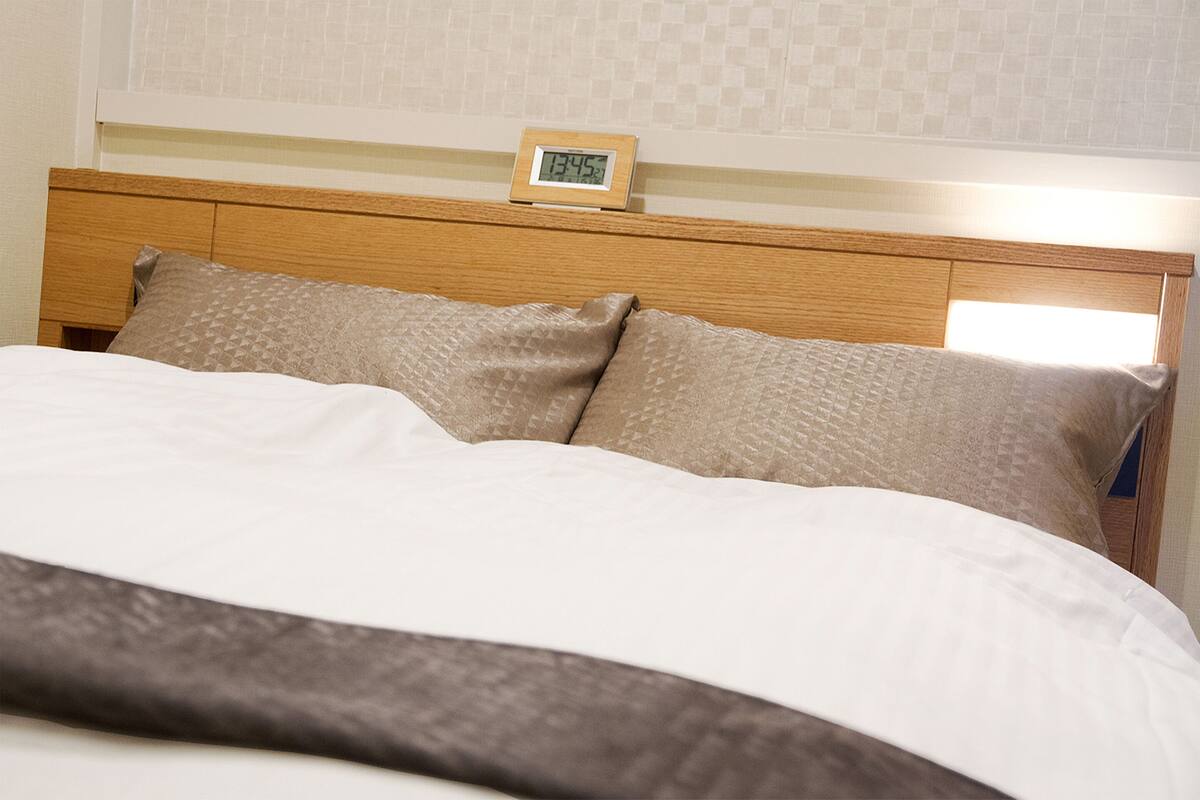 Cama bem arrumada com cabeceira de madeira, duas almofadas e um relógio digital mostrando as horas.