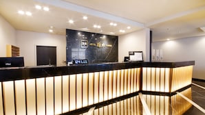 Un mostrador de recepción de hotel con un reloj digital que marca las 12:41, una barra de recepción y una pared con el nombre y el logotipo del hotel.