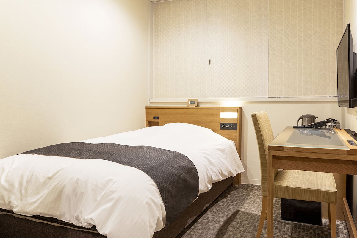 Quarto de hotel com cama, escrivaninha, cadeira, televisão e uma pequena mesa com chaleira.