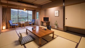A traditional Japanese room with tatami flooring, a wooden table, two blue chairs, a television, and a view of a forested hill. - Hanamaki Onsen Hotel Senshukaku (Hanamaki)